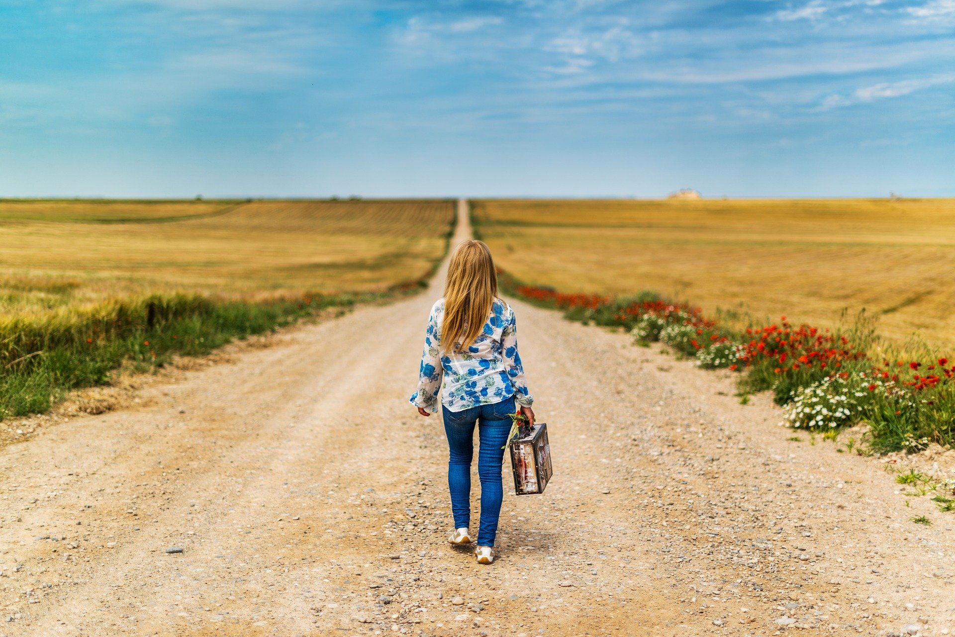 A woman walks down a long dirt road with a field on either side