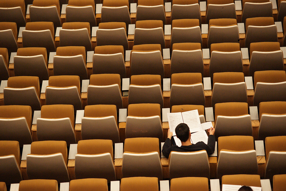 A man sits in an empty classroom of brown seats while he studies the pages in his lap.