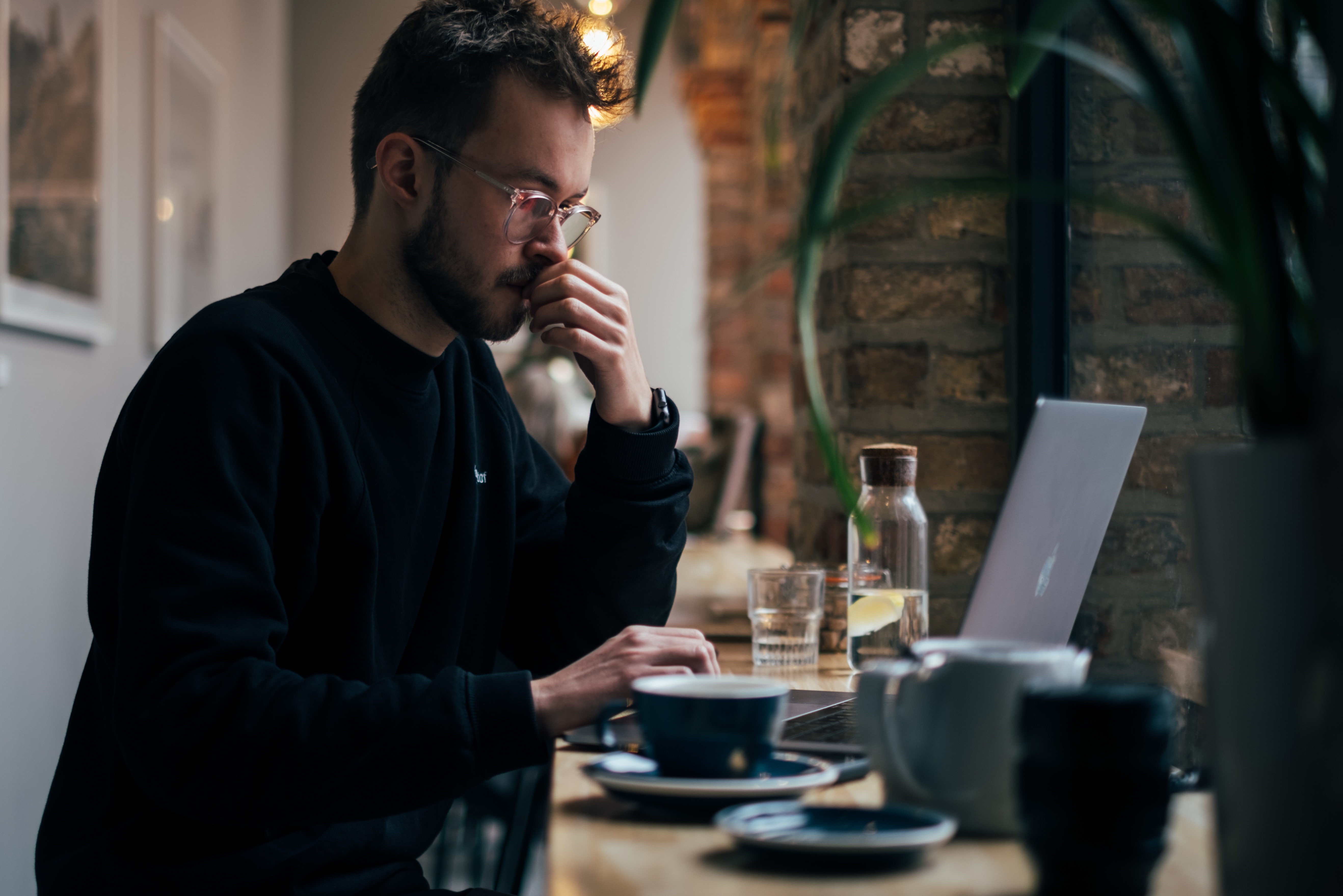 A man stands in front of his open laptop at a coffee shop, as he is deep in thought.