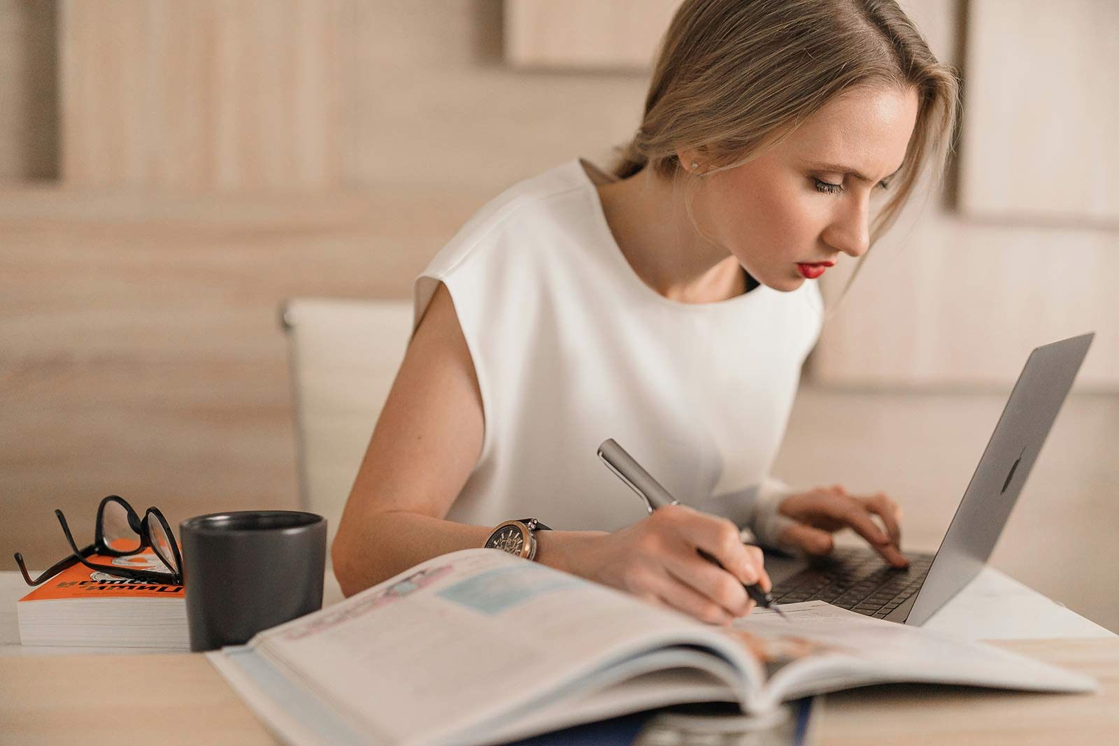 A woman in a white shirt hunches over a laptop as she holds a pen to a textbook.