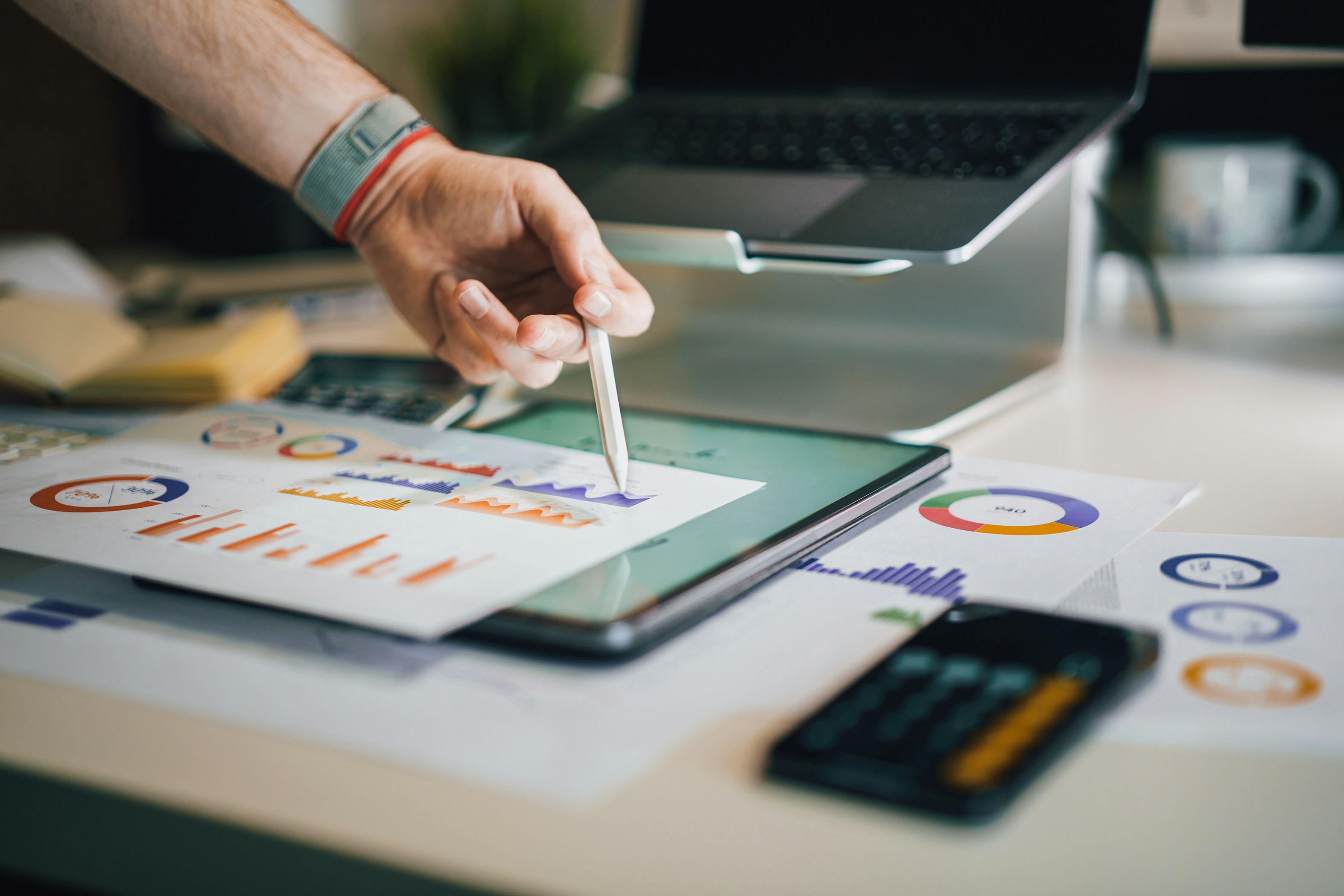 A hand holding a pencil points to graphs and data on paper and digitally.