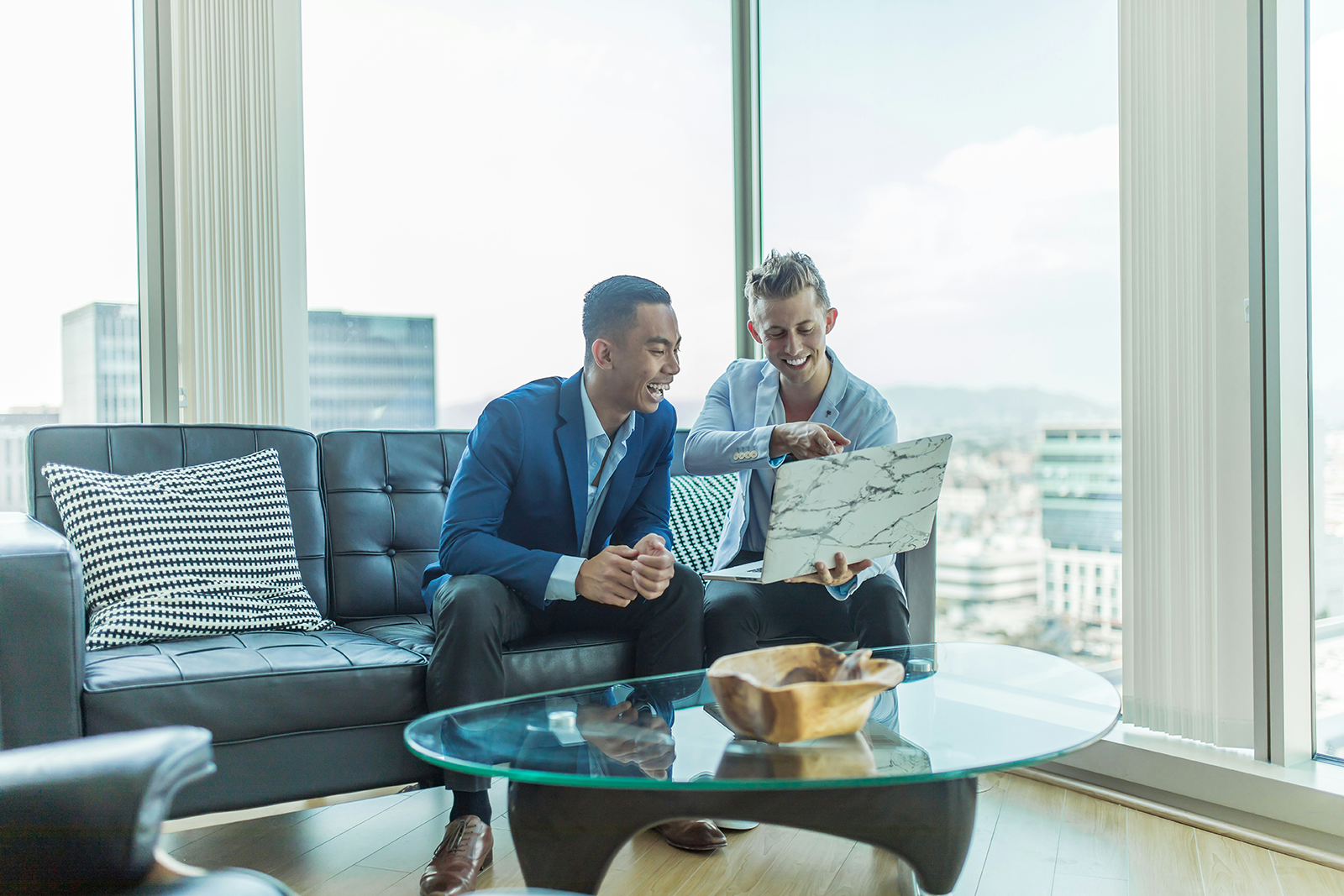 Two men in suits smile as they look at a laptop and sit on a couch.