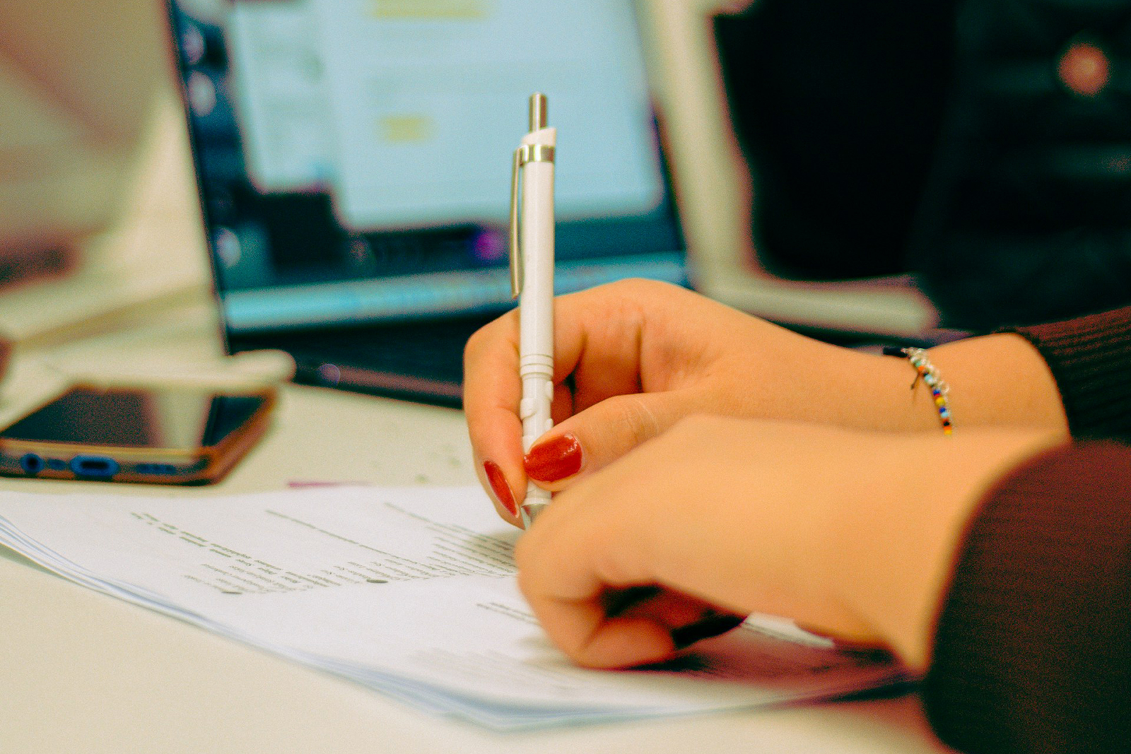 A hand holds a pen and writes on a paper with a laptop in the background.