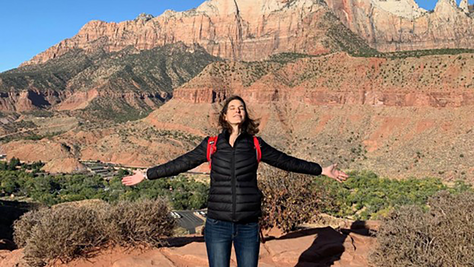 A woman in a black jacket stands in the mountains with arms out wide.